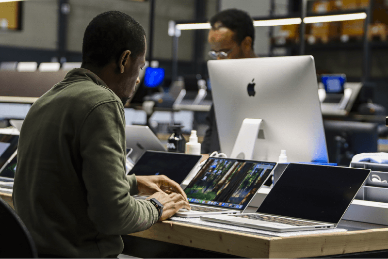 Bovenaanzicht van de SimplyUsed werkplaats met georganiseerde teststations vol MacBooks en iMacs, waar apparaten worden getest.